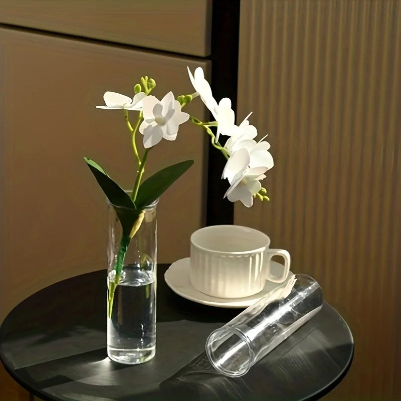 Set de vases cylindriques transparents, trois tailles - Nesty - Vase garni d'orchidées blanches posé sur une table ronde noire près d'une tasse blanche, un second vase couché à côté