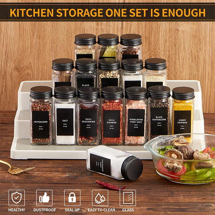 Set de bocaux à épices en verre avec étiquettes Nesty, seize bocaux à épices en verre avec couvercle noir et étiquettes noires (poivre, sel, chili, sel rose, poivre noir, curry), sur présentoir à trois niveaux en bois, bol de salade en fond