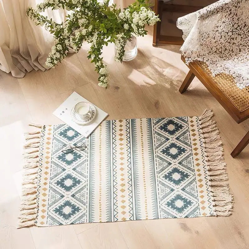 Tapis tissé main en coton et lin Nesty - Franges bohème, variante motif aztèque bleu et doré avec franges, près d'une chaise en rotin et de fleurs