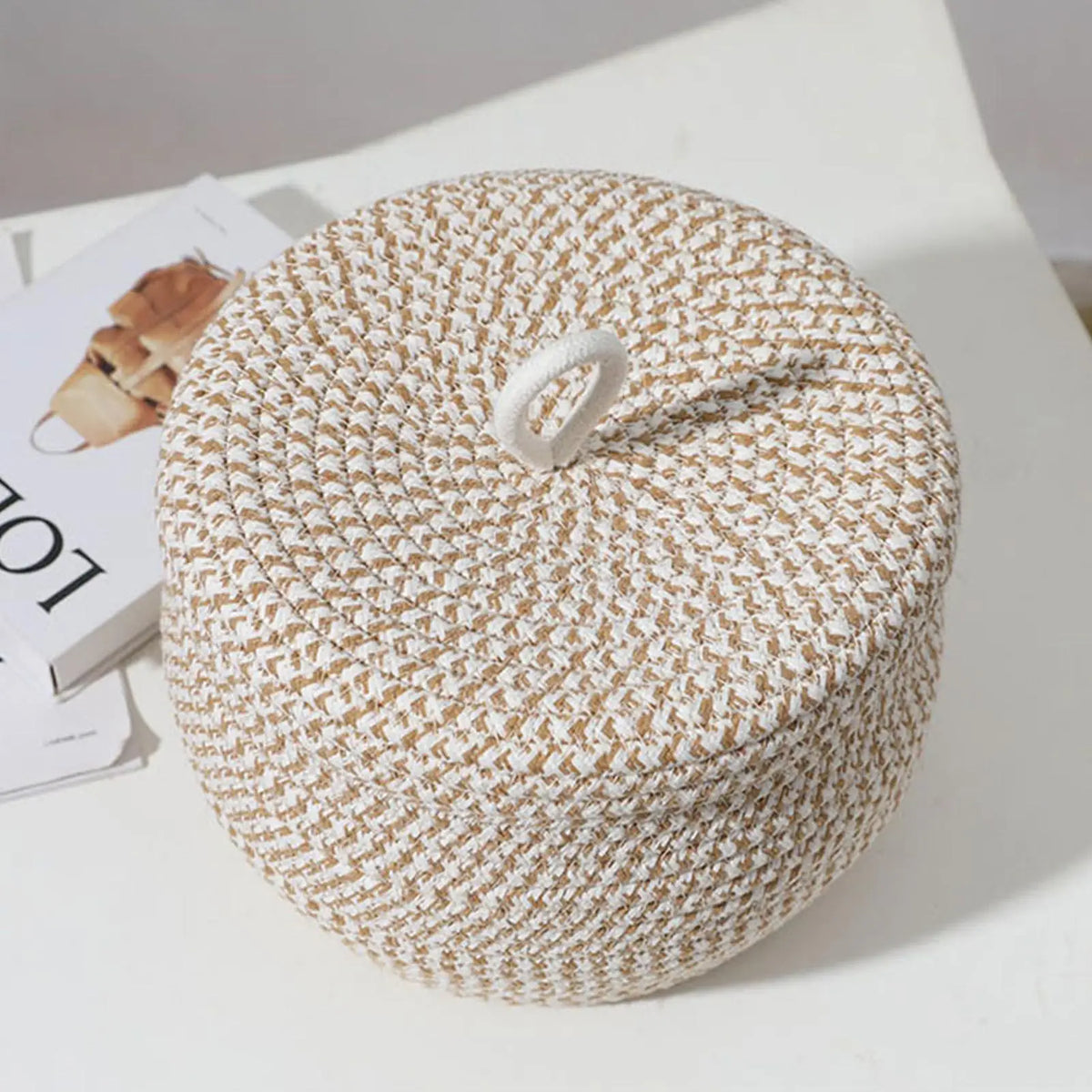 Petit panier rond tressé en corde de coton avec couvercle Nesty, vue de dessus du couvercle fermé avec anse en boucle, sur surface blanche avec magazine