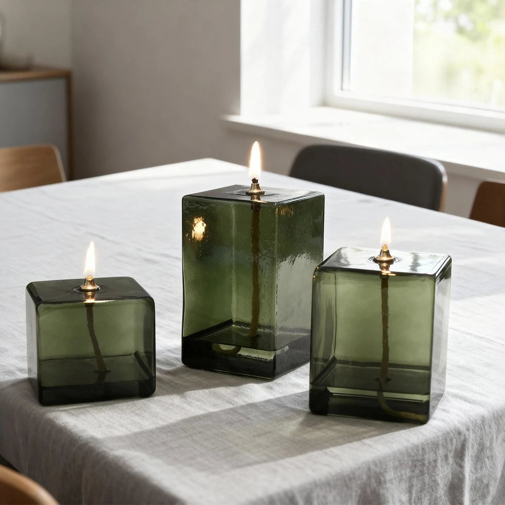 Lampe à huile en verre vert Nesty - Trois lampes cubiques allumées sur une nappe blanche, devant une fenêtre