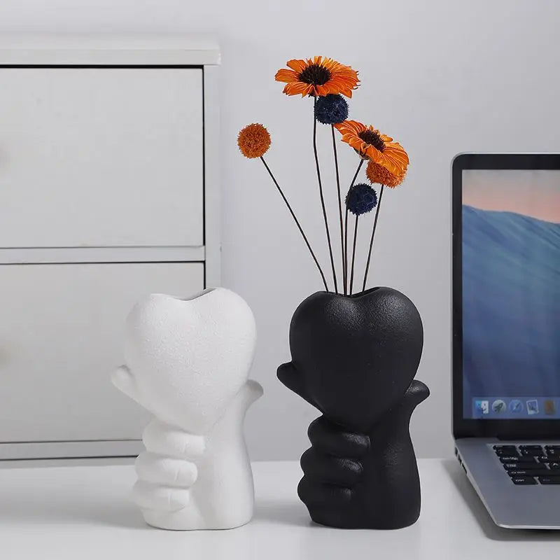 Vase en céramique forme cœur Nesty - Modèles blanc et noir côte à côte, le noir garni de fleurs oranges et bleues, près d'un ordinateur portable