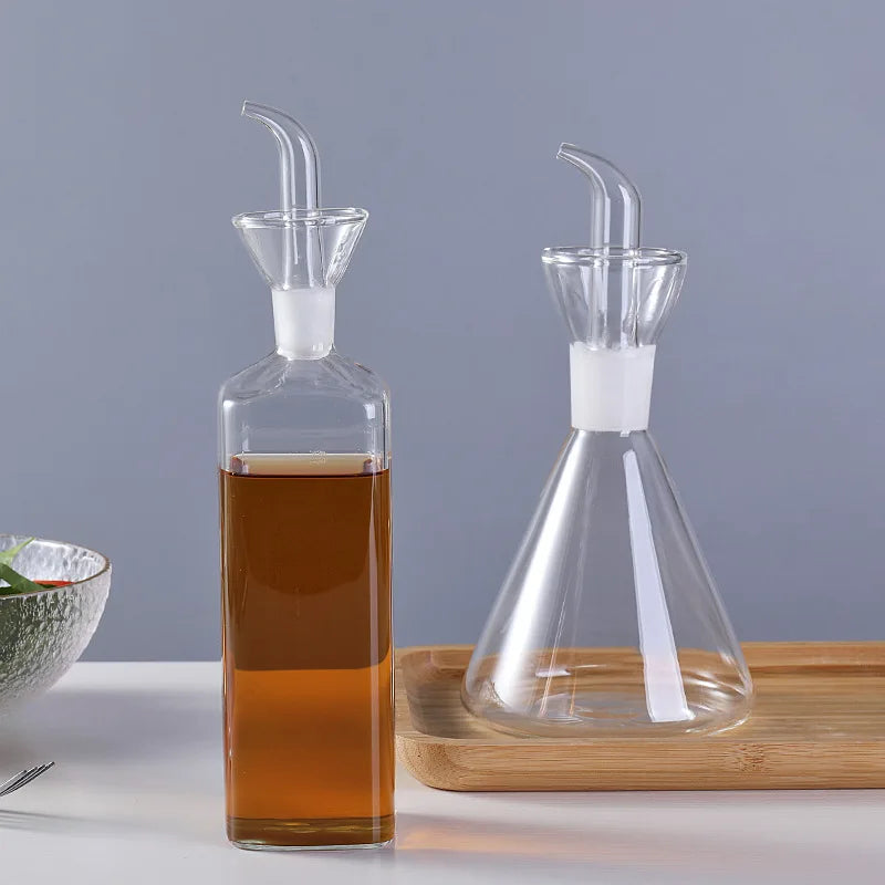 Pot à huile en verre borosilicate, anti-goutte Nesty, bouteille carrée remplie d'huile ambrée à côté d'un modèle conique vide, sur plateau en bambou, fond gris