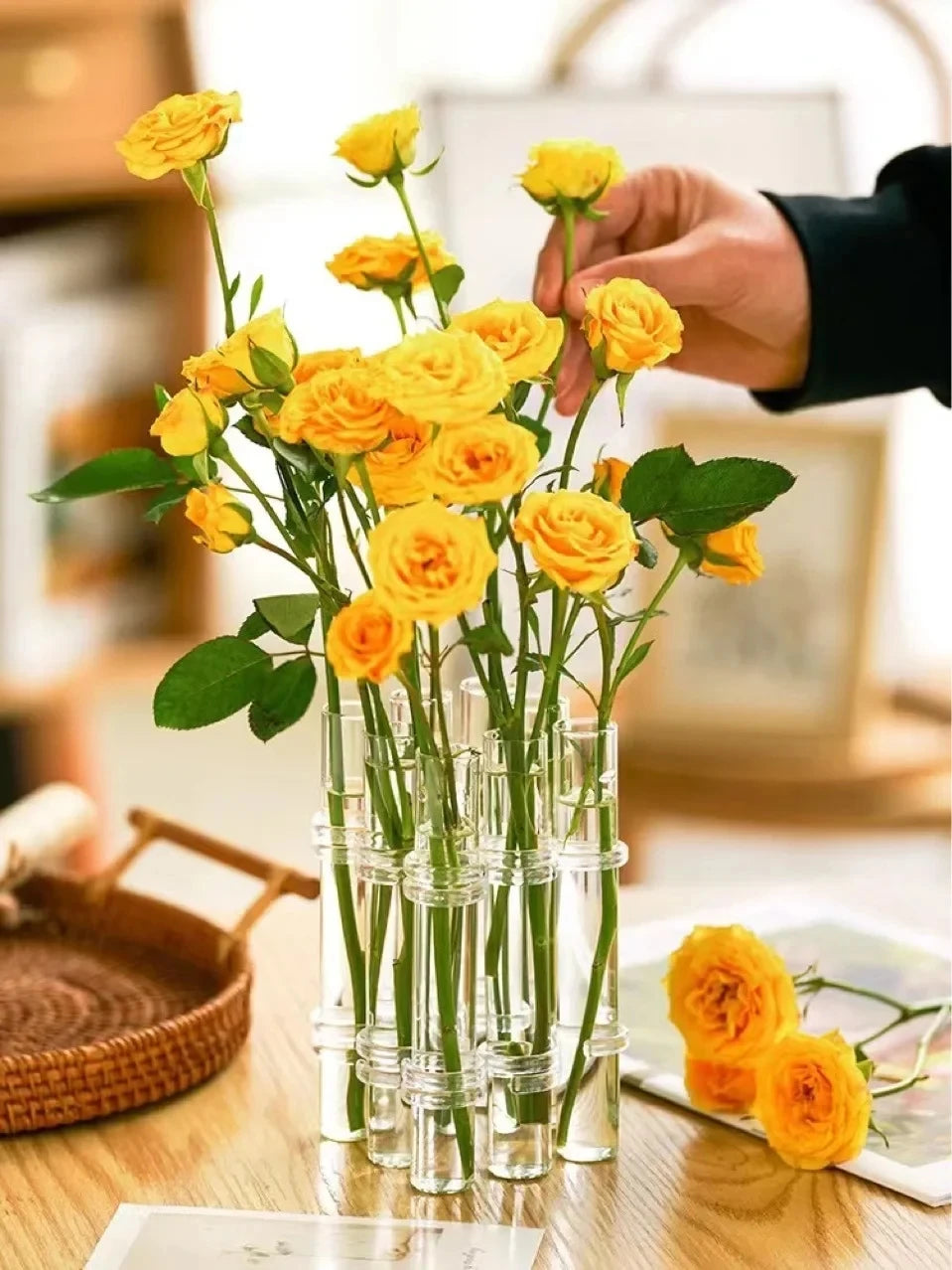 Vase suspendu en verre pour bouturage, lot de tubes transparents Nesty, tubes garnis de roses jaunes, main ajustant une tige, panier en osier à côté