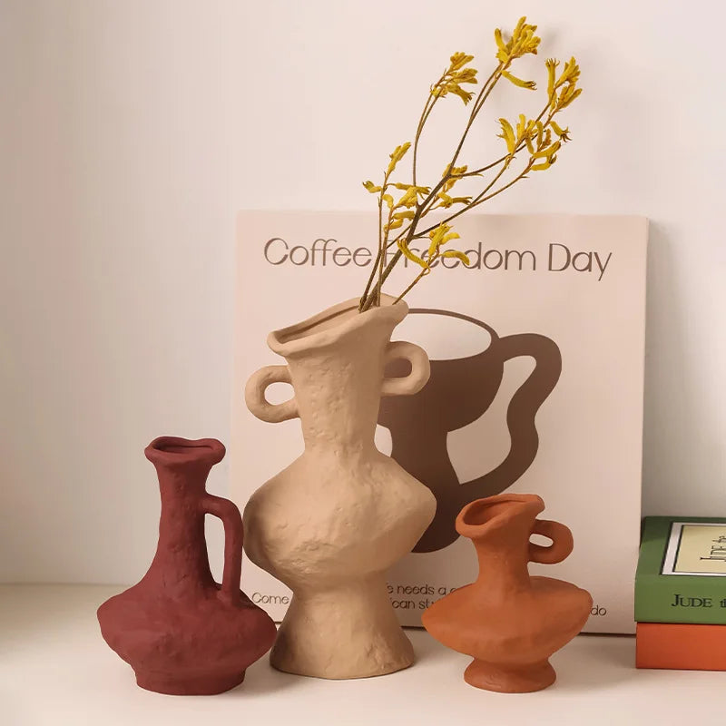 Vase céramique artistique, style nordique rétro - Nesty - Trois vases en terre cuite de formes libres et coloris bordeaux, beige et terracotta, l'un garni de fleurs jaunes, devant une affiche graphique