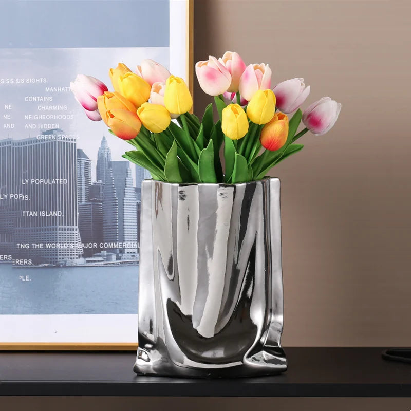 Vase en céramique argentée, forme sac plissé Nesty, effet froissé argenté garni de tulipes multicolores, devant une affiche de ville