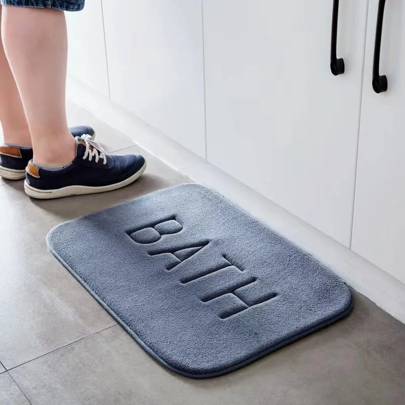 Tapis de bain moelleux en mousse à mémoire de forme - Nesty, variante bleu-gris gaufrée du mot BATH posée au sol près d'un enfant chaussé