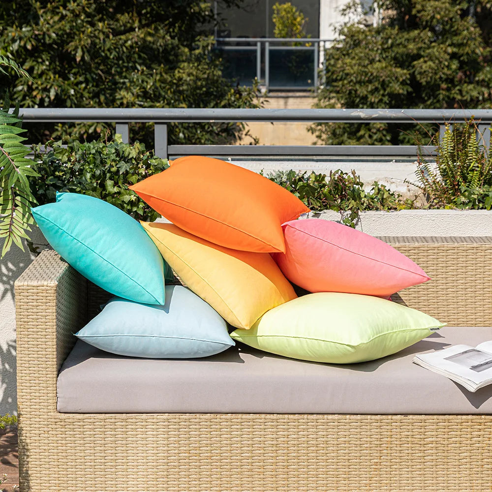 Housses de coussin extérieur imperméables, lot de 2 Nesty, six coussins colorés turquoise, orange, rose, vert clair et bleu clair empilés sur un canapé en rotin extérieur