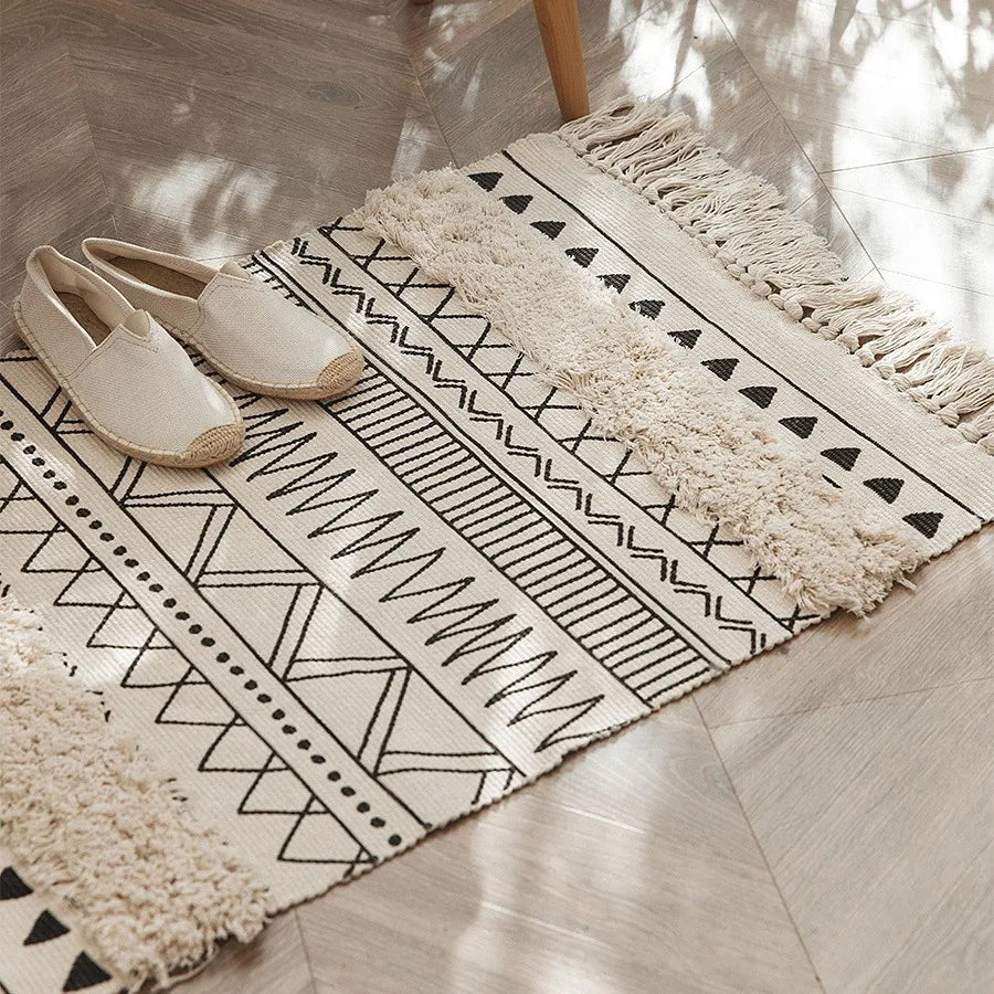 Tapis noué main à franges — motif géométrique - Nesty, variante noir et écru à motifs zigzag et triangles, franges, posée près de chaussures et d'une chaise