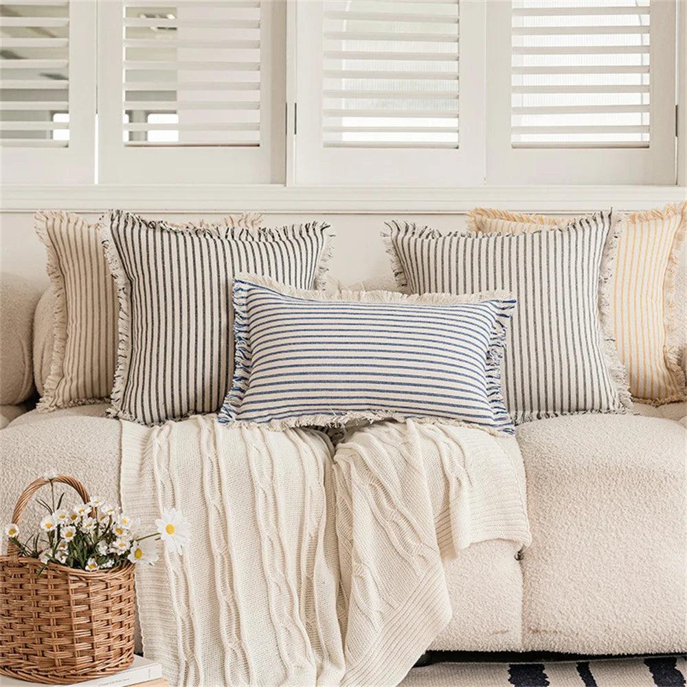 Housse de coussin en lin à rayures, plusieurs formats disponibles Nesty, cinq housses de coussin à rayures (noir, bleu marine, gris, moutarde) avec bordures à franges, posées sur canapé écru et plaid tricoté