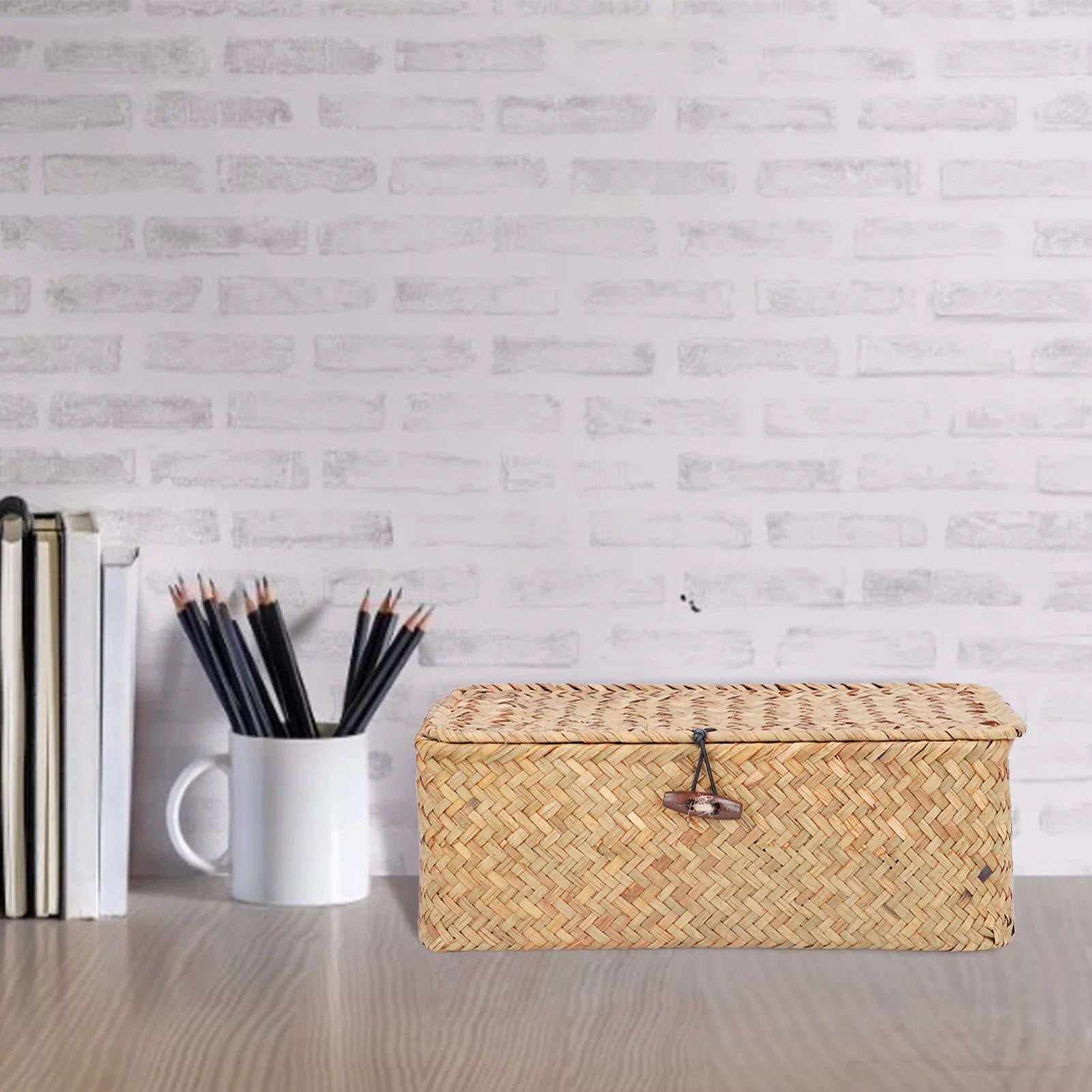 Panier de rangement rectangulaire en jonc de mer tressé avec couvercle Nesty, fermé sur un bureau clair avec pot à crayons et pile de livres, fond blanc à lattes
