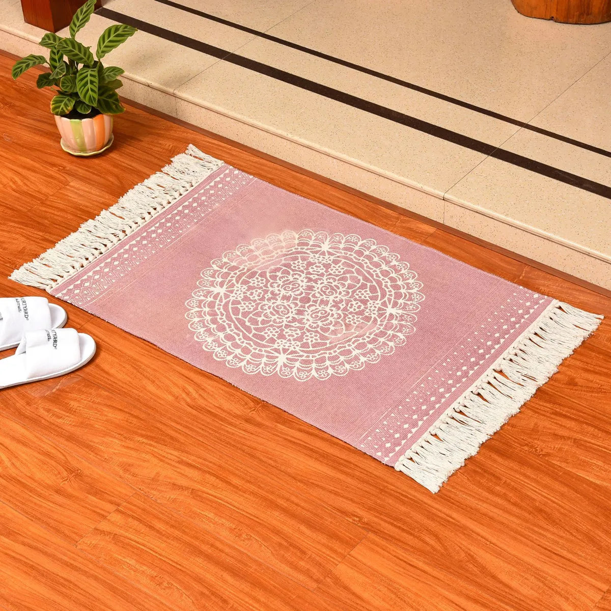 Tapis tissé main en coton et lin Nesty - Franges bohème, variante rose à motif dentelle mandala avec franges, posée près de chaussons et d'une plante