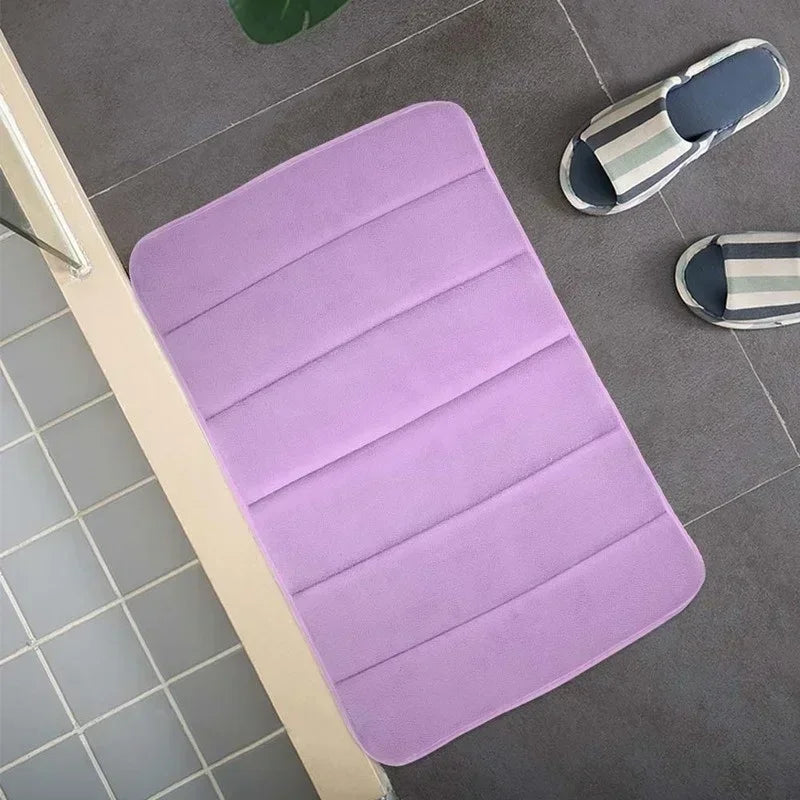 Tapis de bain violet à rainures, posé sur un sol carrelé gris près d'une paire de chaussons rayés.