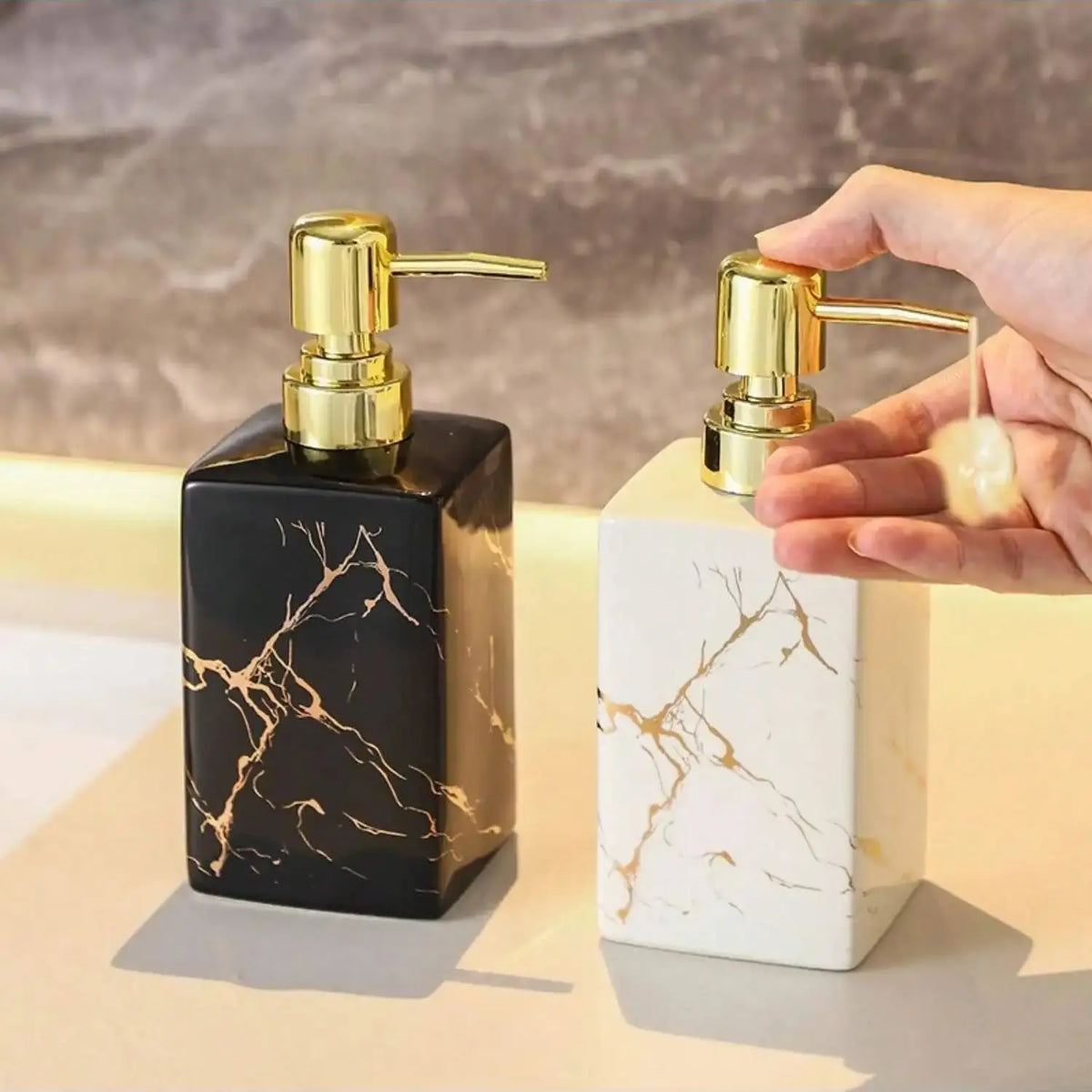 Distributeur de savon en céramique effet marbre Nesty, distributeur de savon céramique marbré noir posé sur vasque, main appuyant sur la pompe dorée du distributeur blanc pour faire couler du savon