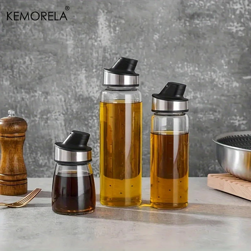 Burette à huile en verre à fermeture automatique Nesty, trois burettes de tailles différentes (huile ambrée et sauce foncée) marque Kemorela, sur plan de travail avec moulin à poivre en bois et poêle