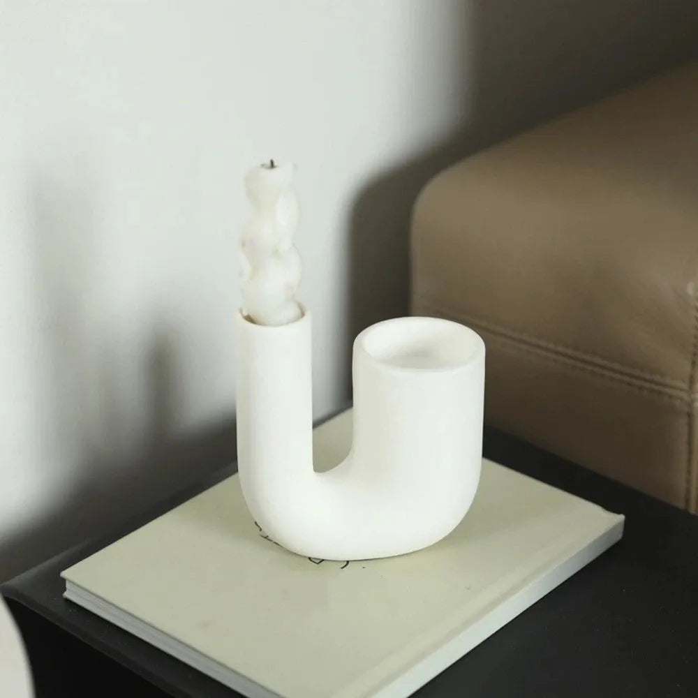 Bougeoir céramique en U finition blanche mate, bougie sculptée en cire, posé sur un carnet près d'un fauteuil en cuir - Nesty