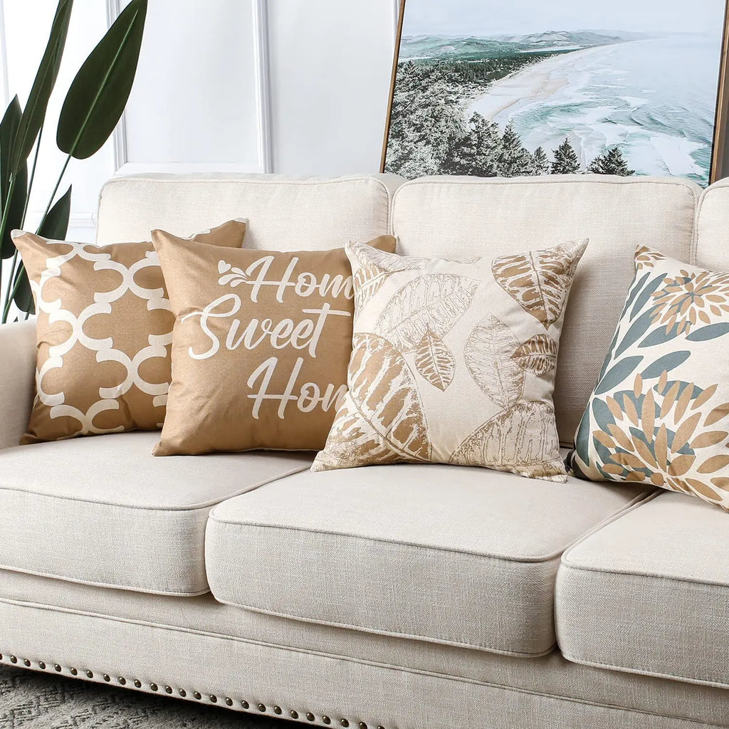 Housse de coussin en lin, motif feuille géométrique brun clair, modèle 2 Nesty, quatre housses de coussin beige et taupe posées sur canapé devant un tableau de paysage côtier (quadrillage doré, "Home Sweet Home", feuilles XL, fleurs bicolores)