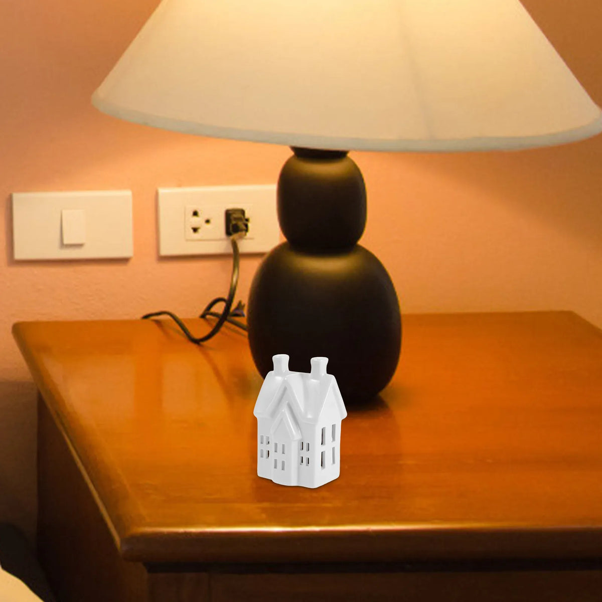 Photophore maisonnette ajourée en céramique Nesty - Maisonnette posée sur une table de chevet en bois devant une lampe noire allumée