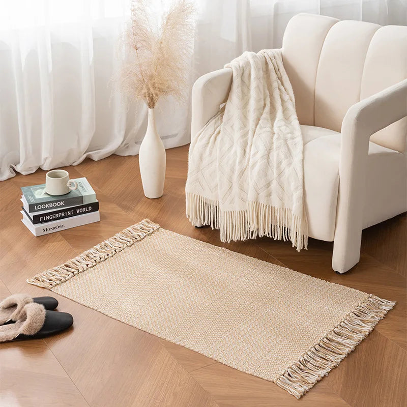 Tapis tressé en coton et lin — franges - Nesty, tapis tressé beige à franges posé près d'un fauteuil crème avec plaid à franges, livres et vase