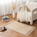 Tapis tressé en coton et lin — franges - Nesty, tapis tressé beige à franges posé près d'un fauteuil crème avec plaid à franges, livres et vase