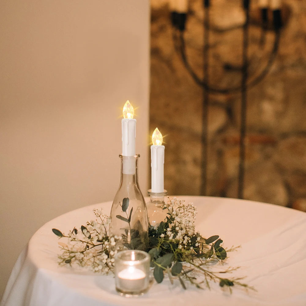 Bougie LED sans flamme, mèche noire, télécommande incluse Nesty, deux bougies LED plantées dans des bouteilles en verre entourées de gypsophile, bougie votive au premier plan, sur table dressée