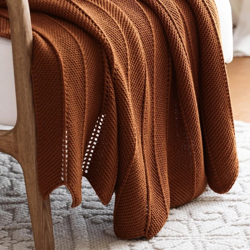 Plaid tricoté en acrylique coloris brun rouille, à mailles ajourées, posé sur un fauteuil en bois près d'un tapis à motifs - Nesty