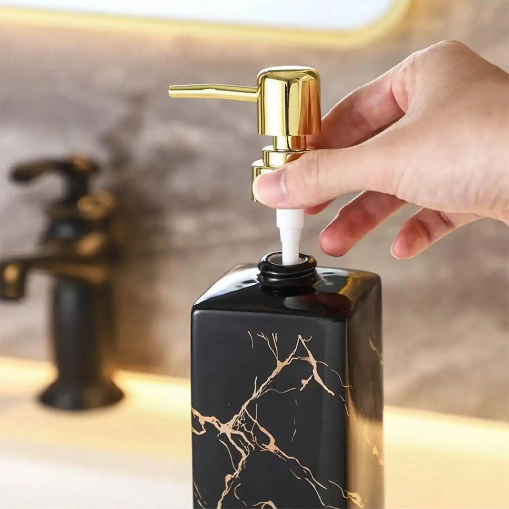 Distributeur de savon en céramique effet marbre Nesty, gros plan sur une main insérant le tube de pompe dorée dans le col d'un distributeur de savon céramique marbré noir