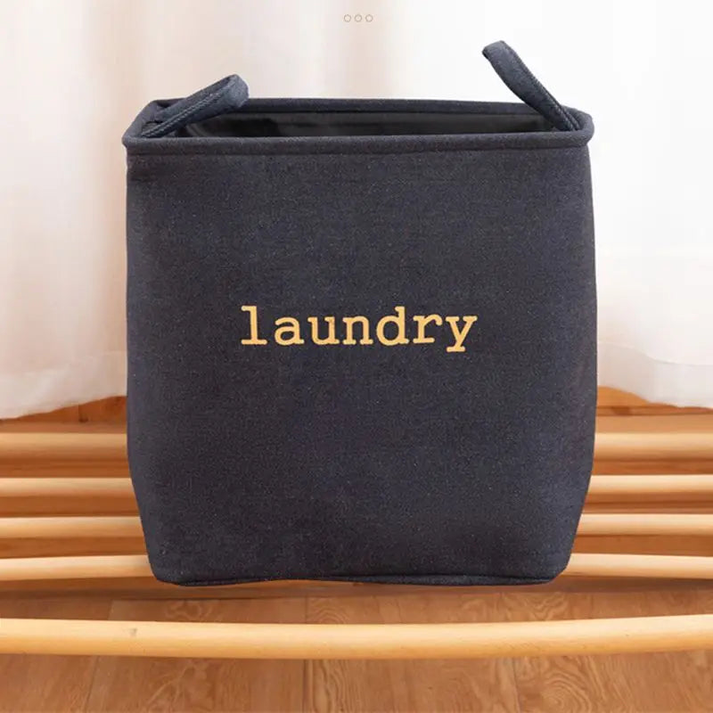 Panier à linge pliable en toile de jute Nesty - Coloris noir avec inscription laundry dorée, posé sur banc en bois