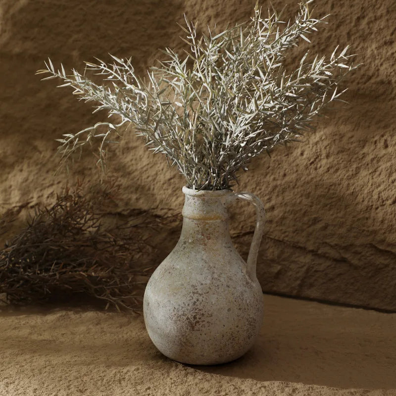 Vase en céramique style wabi-sabi pour fleurs séchées Nesty, cruche en céramique effet vieilli avec anse, garnie de branches d'olivier séchées argentées, devant un mur en pierre