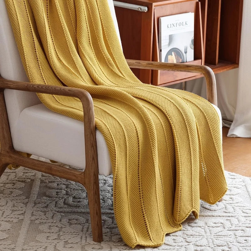 Plaid tricoté en acrylique coloris jaune moutarde, posé sur un fauteuil en bois clair près d'un meuble bibliothèque - Nesty