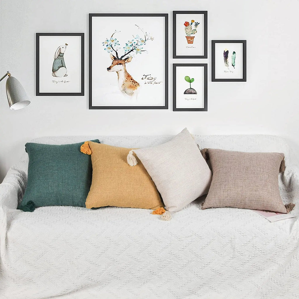 Housse de coussin en lin avec pompons, toutes saisons Nesty, quatre housses de coussin à pompons colorés (vert sapin, moutarde, écru, taupe) posées sur canapé blanc sous une galerie de cadres illustrés