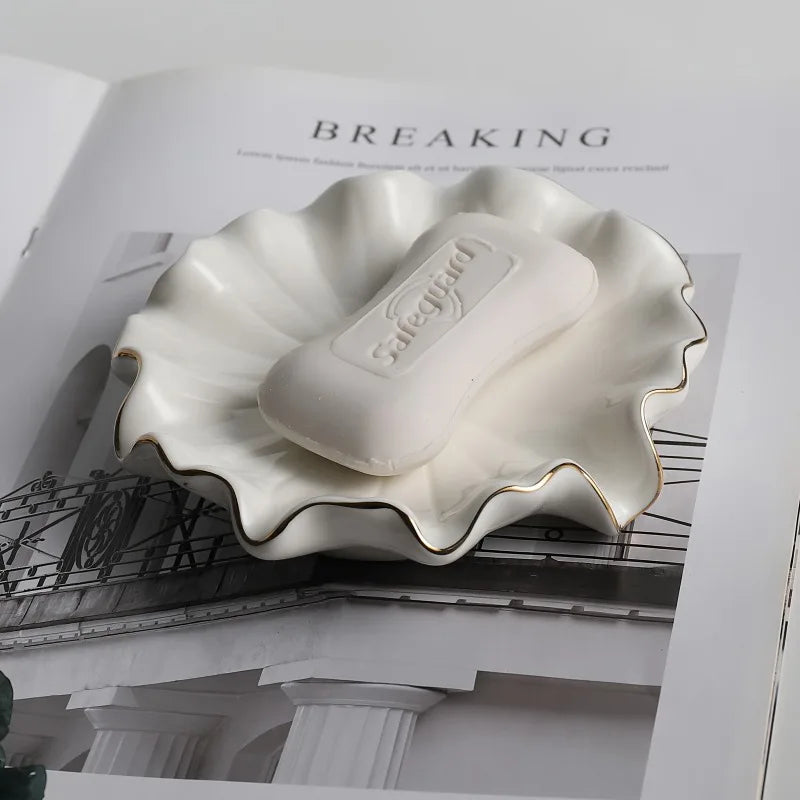 Porte-savon en céramique blanche forme coquillage à liseré doré avec savon, posé sur un magazine ouvert - Nesty