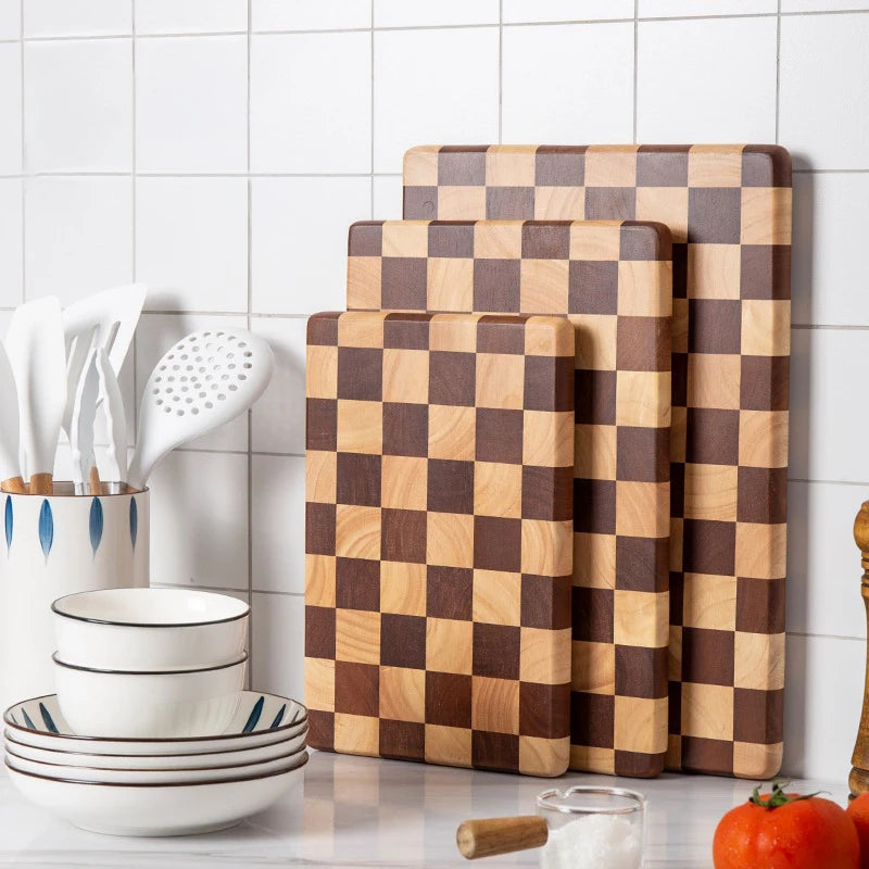 Planche à découper en bois massif, motif mosaïque Nesty, trois tailles de planches à damier bicolore appuyées contre un mur carrelé blanc, avec ustensiles de cuisine et bols