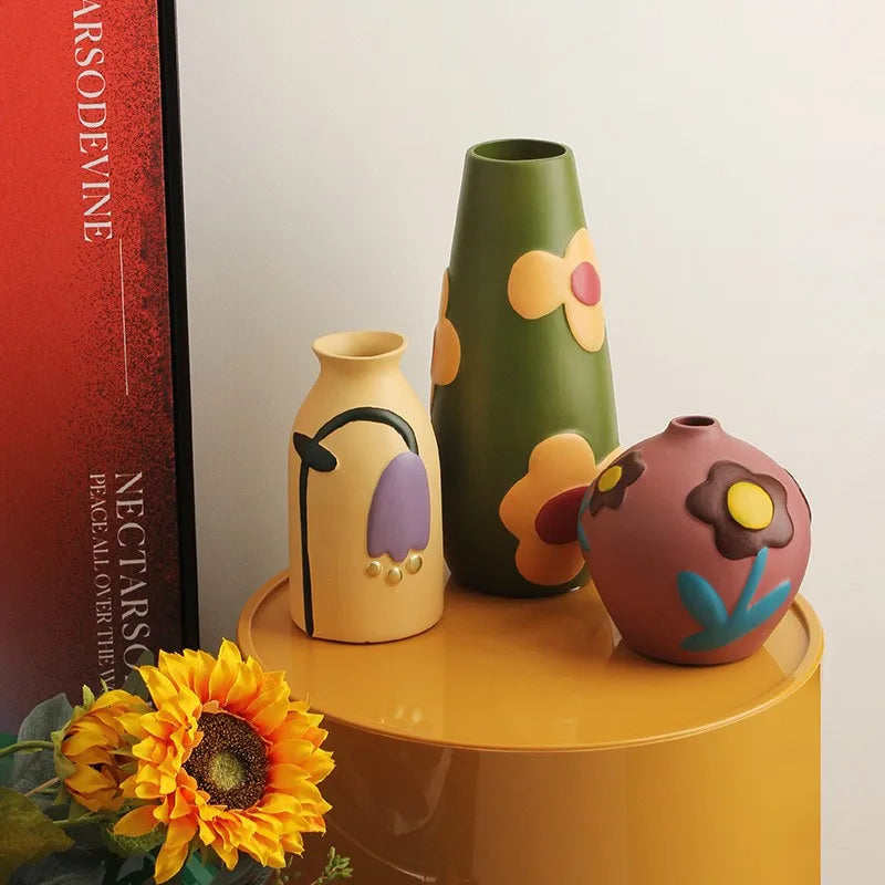 Vase céramique émaillé teintes Morandi, style nordique - Nesty - Trois vases peints beige à motif floral mauve, vert olive à fleurs et marron-rose à fleur, sur une table ronde jaune avec tournesols