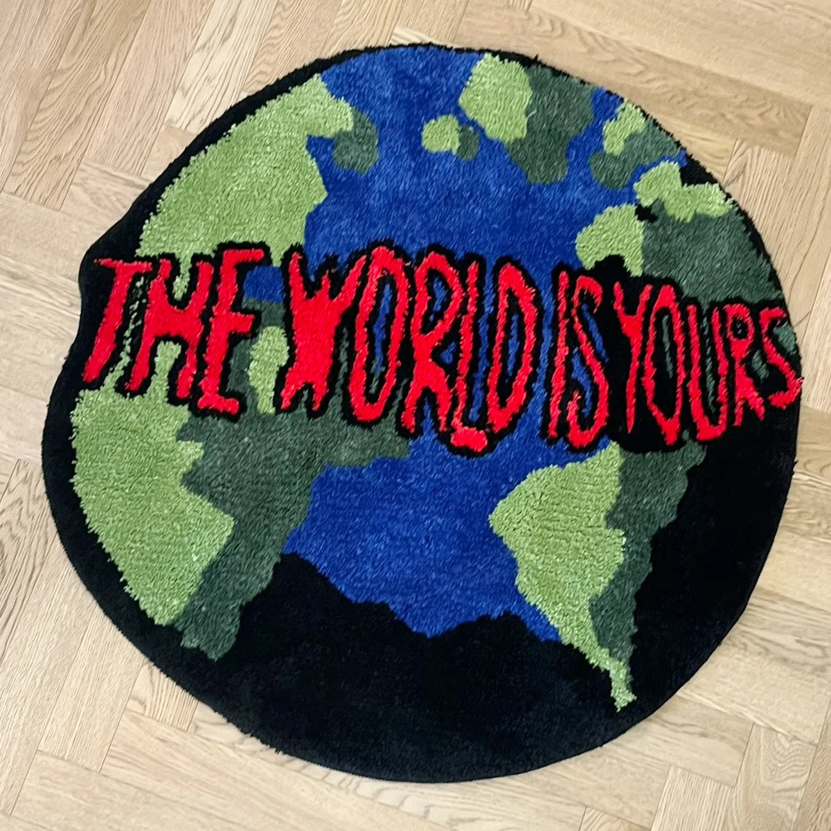 Tapis rond en acrylique — motif animalier - Nesty, tapis rond noir façon globe terrestre avec l'inscription The World Is Yours en rouge, posé sur un parquet