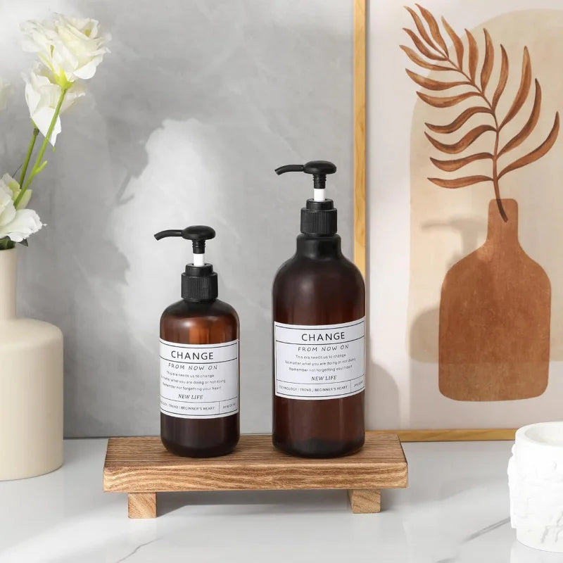 Piédestal en bois Nesty - Présentoir pour savon ou bougie, piédestal en bois supportant deux flacons pompe ambrés étiquetés Change, près d'un vase de fleurs blanches