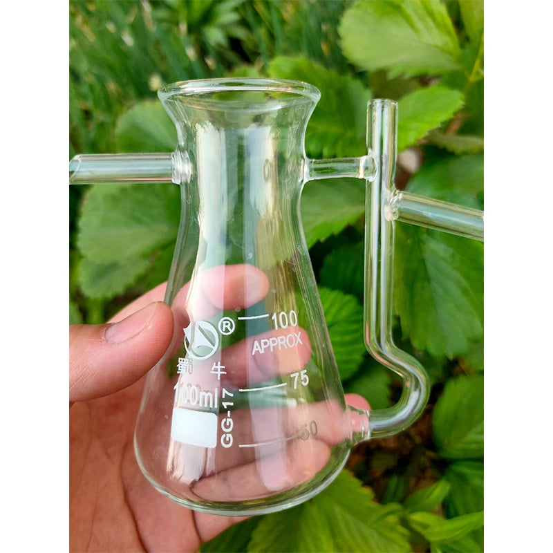 Main tenant une fiole en verre gradué avec tubes de distillation, devant un feuillage vert - Nesty