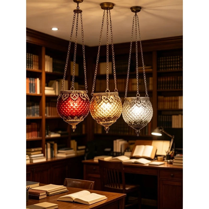 Trio de photophores suspendus en verre Nesty - Photophores rouge, doré et argenté allumés, suspendus au plafond d'une bibliothèque avec bureau et livres