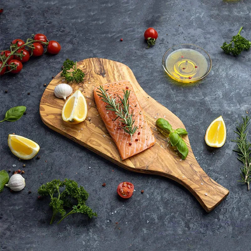 Planche à découper en bois d'olivier, forme irrégulière Nesty, planche irrégulière garnie d'un filet de saumon au romarin, citron, tomates et ail, bol d'huile à côté, fond sombre