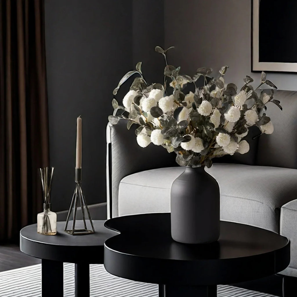 Vase en métal noir mat, finition sablée Nesty, cylindrique garni d'un bouquet de boules blanches et eucalyptus argenté, sur table noire dans un salon sombre