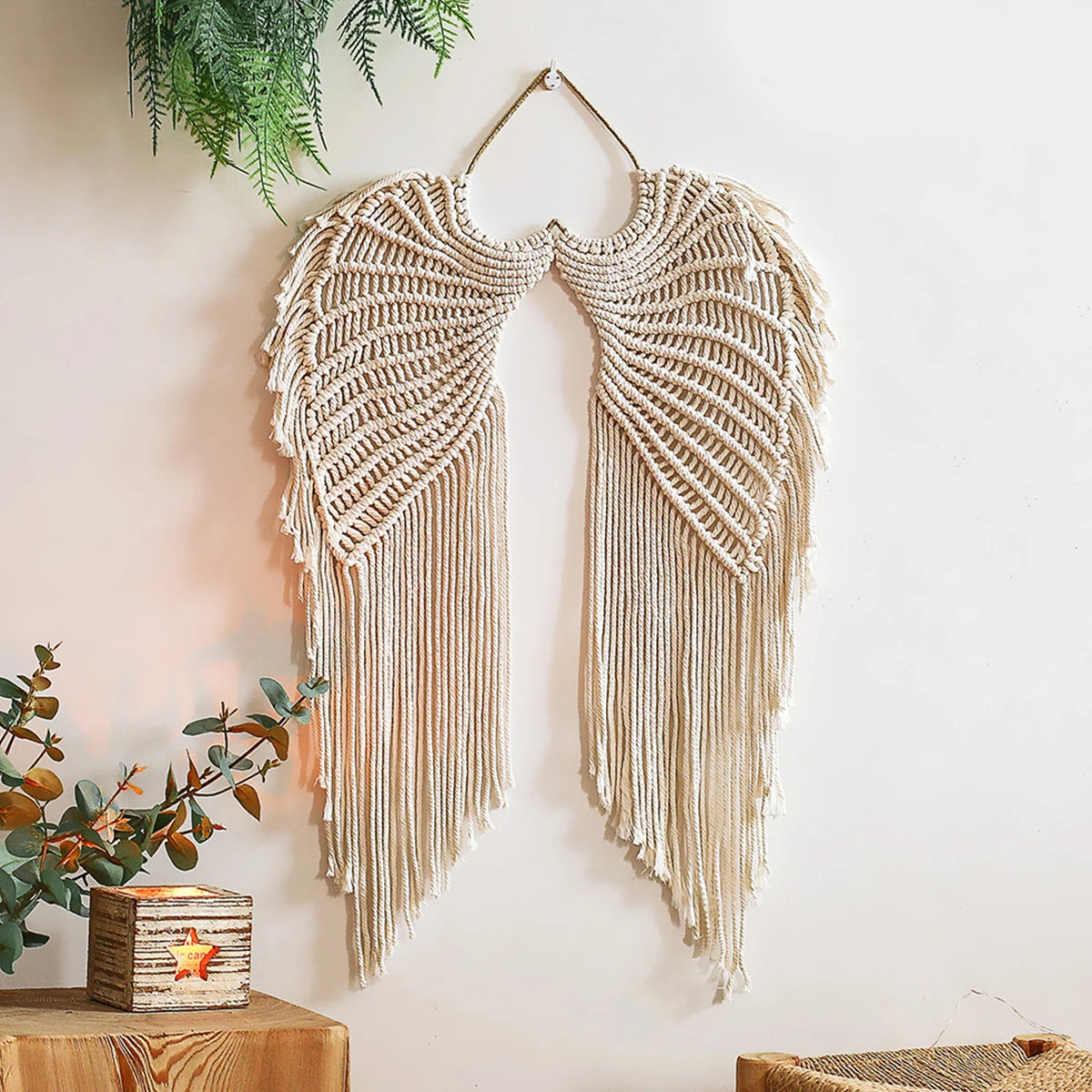 Tenture murale tissée à forme d'ailes Nesty, ailes en macramé beige aux longues franges, suspendues sur un mur blanc avec des plantes vertes en haut