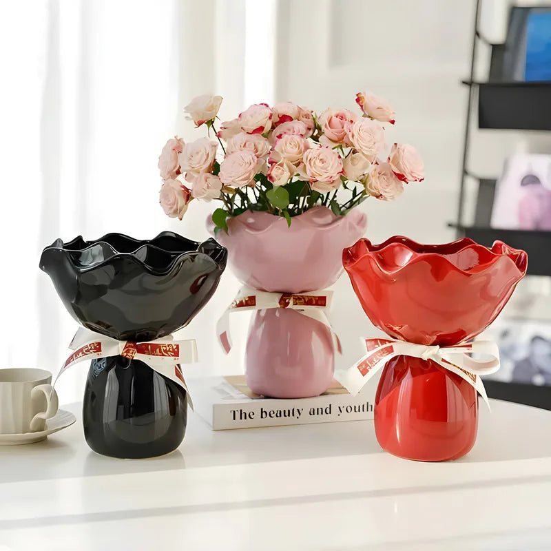 Vase en céramique en forme de rose, modèle bureau Nesty, trois vases céramique à col ondulé noué de ruban, coloris noir, rose et rouge, l'un garni de roses roses, posés sur un livre