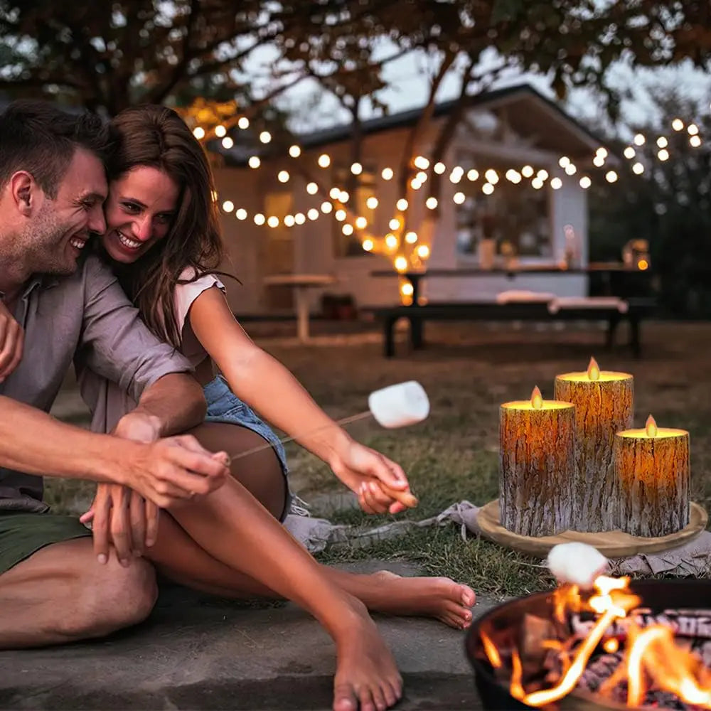 Bougie LED en cire véritable, sans flamme, télécommande incluse Nesty, trois bougies LED effet écorce allumées sur plateau en bois, couple assis autour d'un feu de camp en extérieur