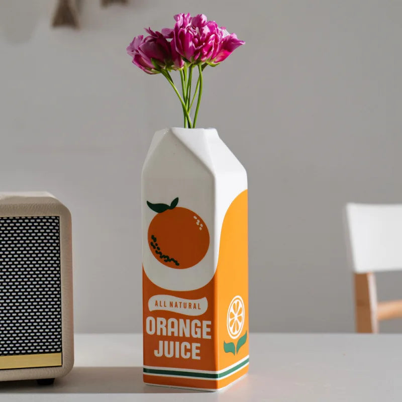 Vase en céramique forme fruit, orange décorative Nesty, vase céramique en forme de brique de jus imprimée "Orange Juice", garni de fleurs roses, sur table blanche