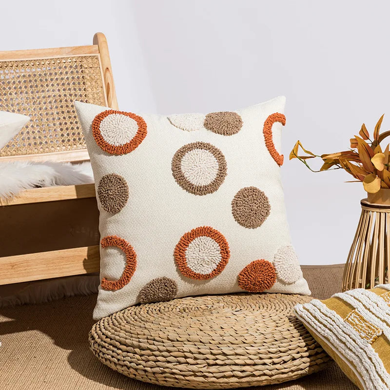 Housse de coussin bouclette à broderie géométrique bohème Nesty, coussin blanc à motif de ronds bruns et orange en bouclette, posé sur une chaise en rotin