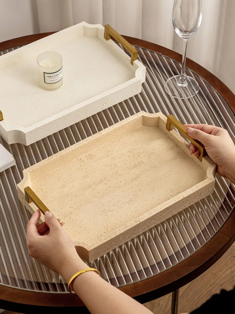 Duo de plateaux en travertin à poignées dorées, mains soulevant le plateau beige, verre à pied en arrière-plan - Nesty