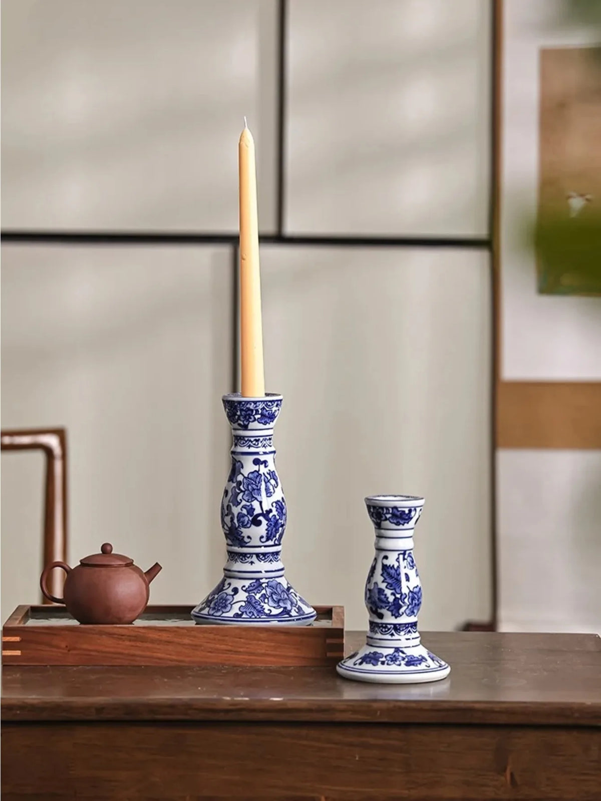 Bougeoir en porcelaine bleue et blanche Nesty - Deux bougeoirs de tailles différentes avec bougie dorée allumée, théière en terre cuite et fenêtre en arrière-plan