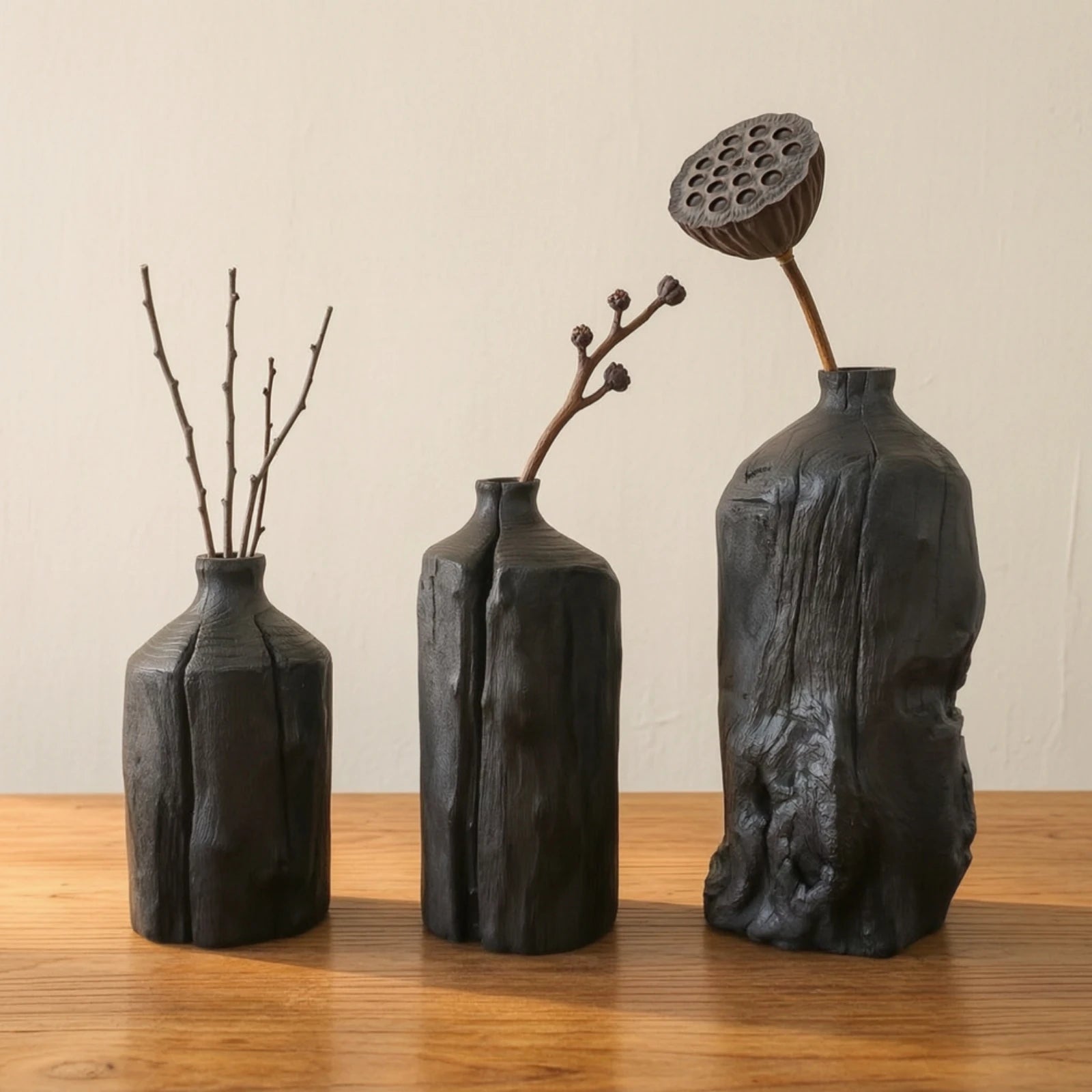 Trio de vases en bois sculpté, style wabi-sabi Nesty, trois tailles noires texturées garnies de brindilles et de graines de lotus séchées, sur table en bois
