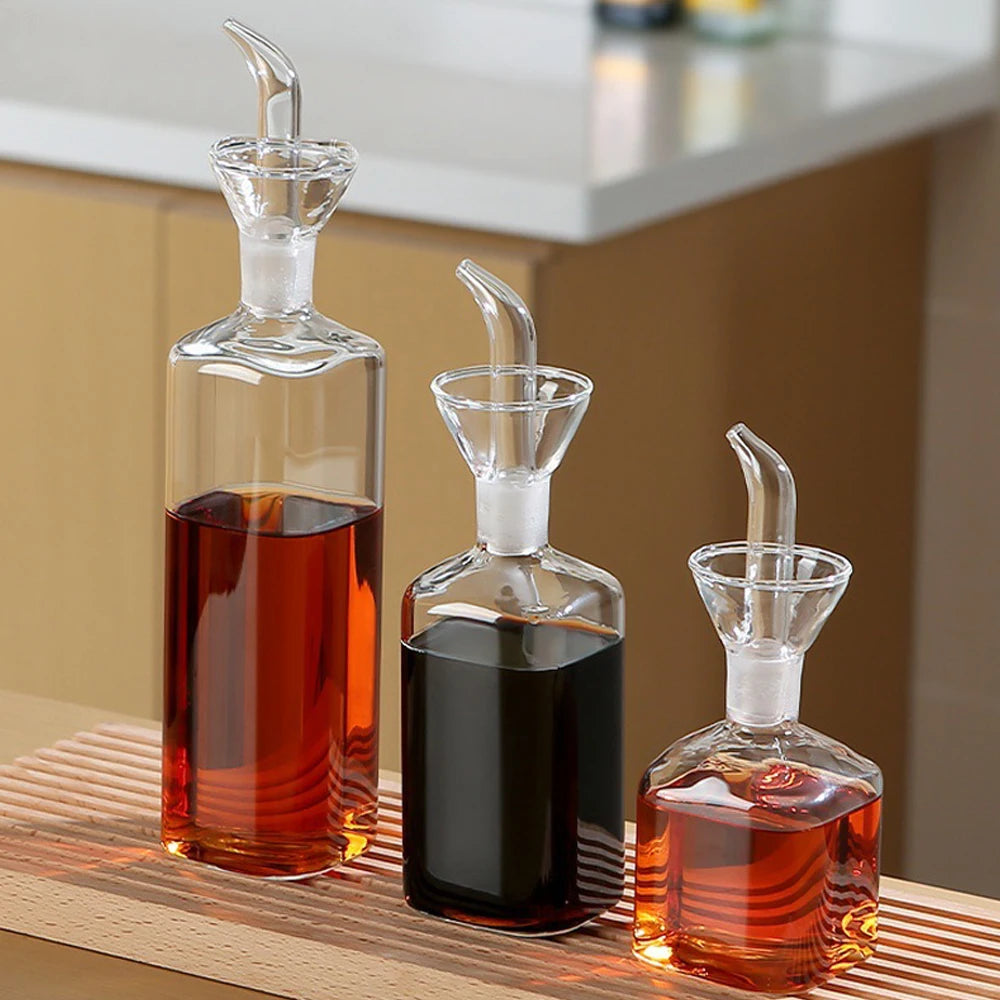 Burette à huile d'olive en verre borosilicate, anti-goutte Nesty, trois burettes en verre de tailles différentes avec bec verseur et bouchon, remplies d'huile ambrée et de sauce foncée, sur plan de travail en bois
