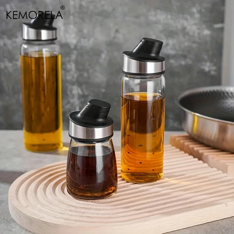 Burette à huile en verre à fermeture automatique Nesty, trois burettes de tailles différentes groupées sur une planche en bois, poêle en arrière-plan